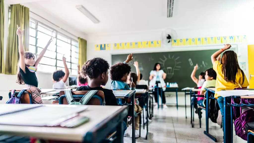 Sala de aula com crianças com as mãos levantadas. Ao fundo, professora na frente da lousa.